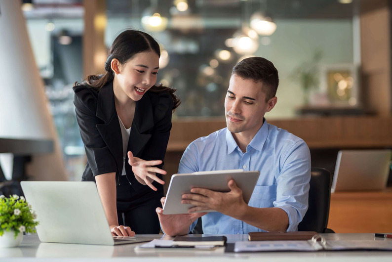 Coworkers Discuss Work Project Over Tablet And Laptop At Desk Scaled